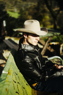 A man wearing a beige cowboy hat and a black leather jacket sits on a green bench in an outdoor setting with warm sunlight and a blurred background.