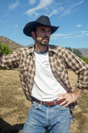 Man wearing a cowboy hat, plaid shirt, and jeans standing outdoors with mountains in the background.