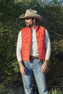 Man wearing a cowboy hat, red vest, and jeans standing in a natural setting with trees and rocks.