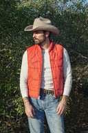 Man wearing a cowboy hat, red vest, and jeans standing in a natural setting with trees and rocks.