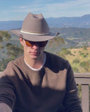 Man in sunglasses and a brown sweater wearing the Matty wool felt cowboy hat in Grey, with hills behind him.